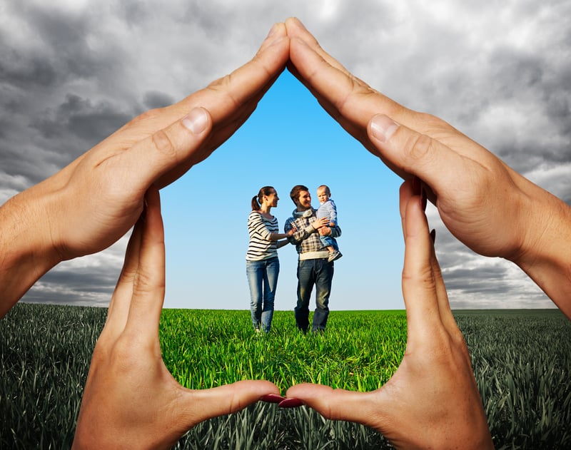 Family of three in sunny field protect to by hands in the shape of house